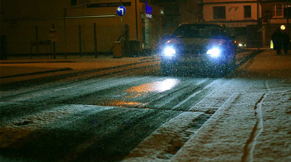 A car on a snowy road in the night, headlights illuminating the snowfall